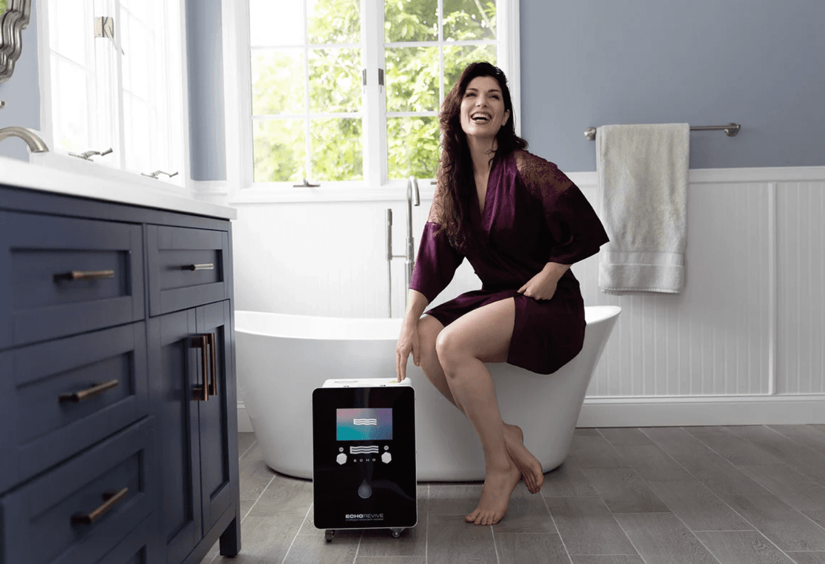 Woman in spa robe using Echo Revive Bath Machine - Hydrogen Water | 1.6 ppm, Lab Tested beside a bathtub, promoting wellness and hydration benefits.
